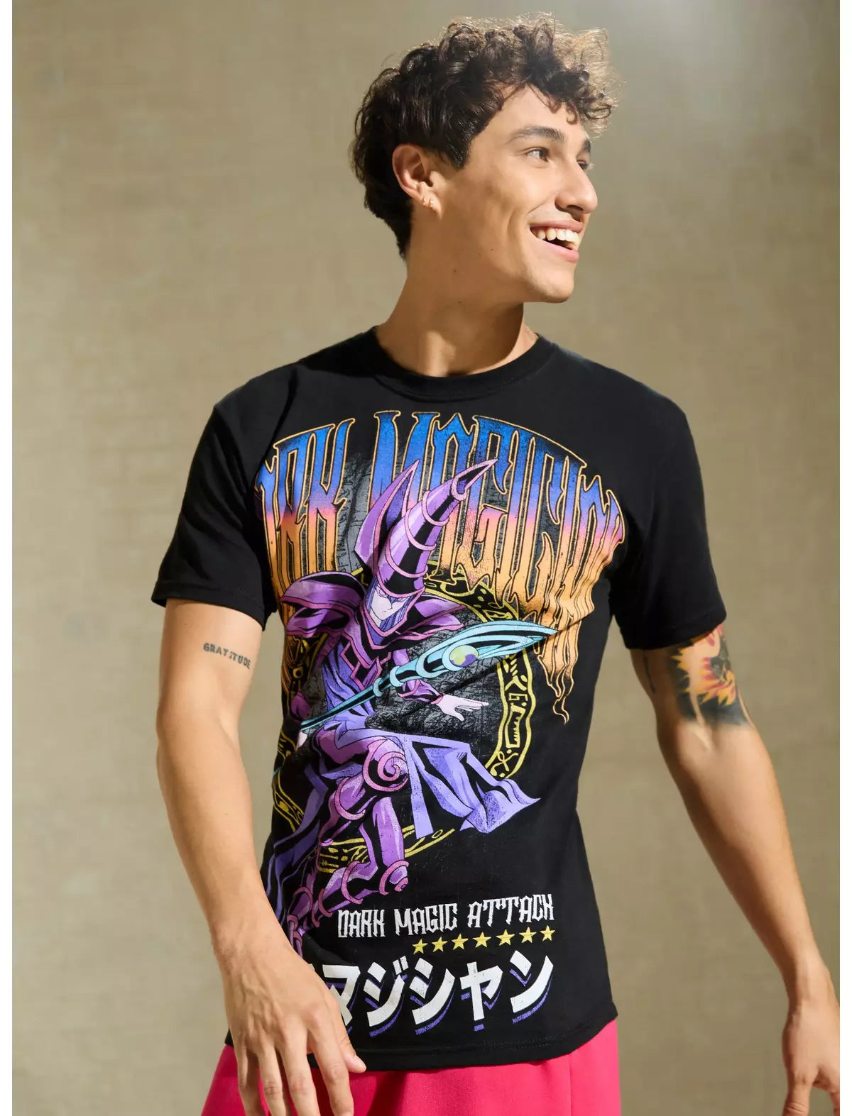 A man wearing a black graphic t-shirt with a colorful 'Dark Magic Attack' print featuring a purple wizard and Japanese text, paired with pink shorts, set against a textured beige background.