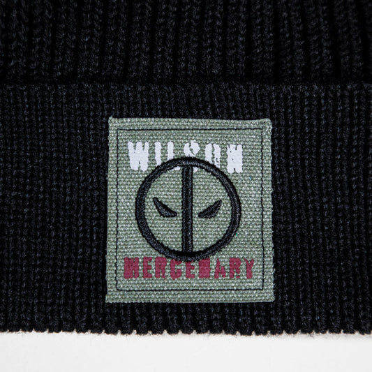 Close-up of a black ribbed knit beanie featuring a square woven patch on the cuff. The patch is sage green with a black circular face emblem, white 'WILSON' text at the top, and pink 'MERCENARY' text at the bottom.