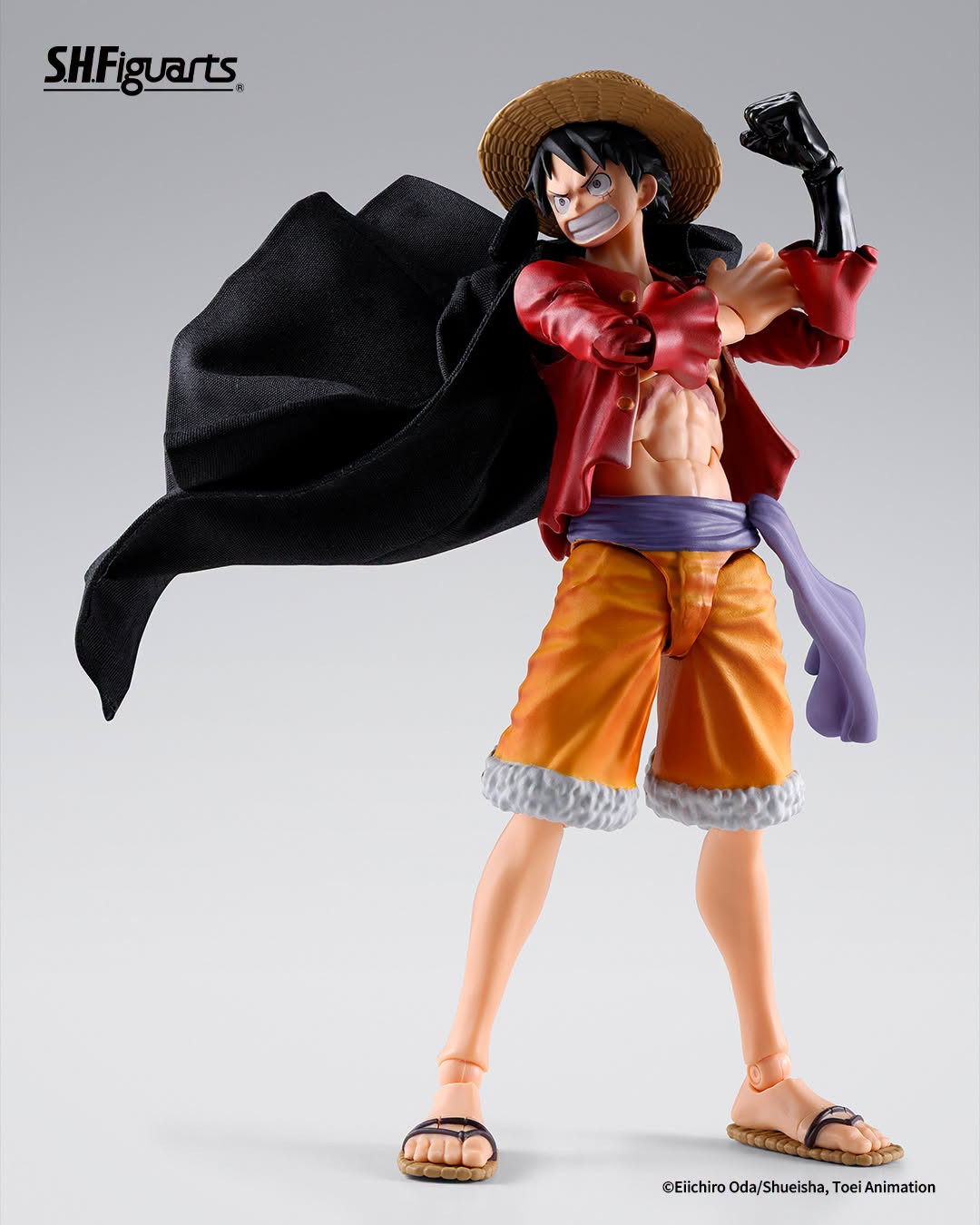 SH Figuarts One Piece Luffy action figure in a dynamic pose, wearing his signature straw hat, red jacket, orange shorts, and black cape against a light gray studio background.