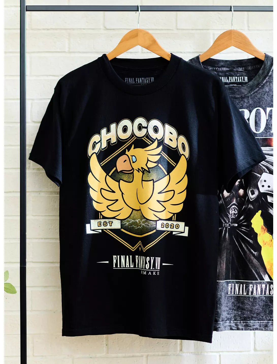 A black short-sleeve graphic T-shirt hanging on a wooden hanger against a light gray background. The shirt features a large gold and beige chakra-style bird graphic with the text 'CHOCOBO' and 'Final fantasy' on the front.