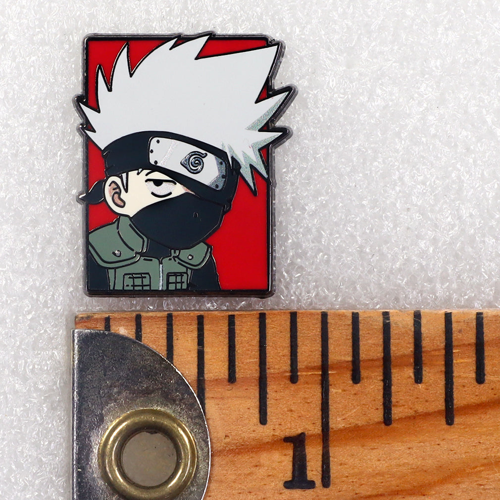 A small enamel pin featuring a stylized anime character with spiky white hair, a black face covering, and a red background, shown next to a wooden ruler for scale on a white textured surface.