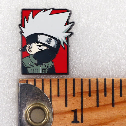 A small enamel pin featuring a stylized anime character with spiky white hair, a black face covering, and a red background, shown next to a wooden ruler for scale on a white textured surface.