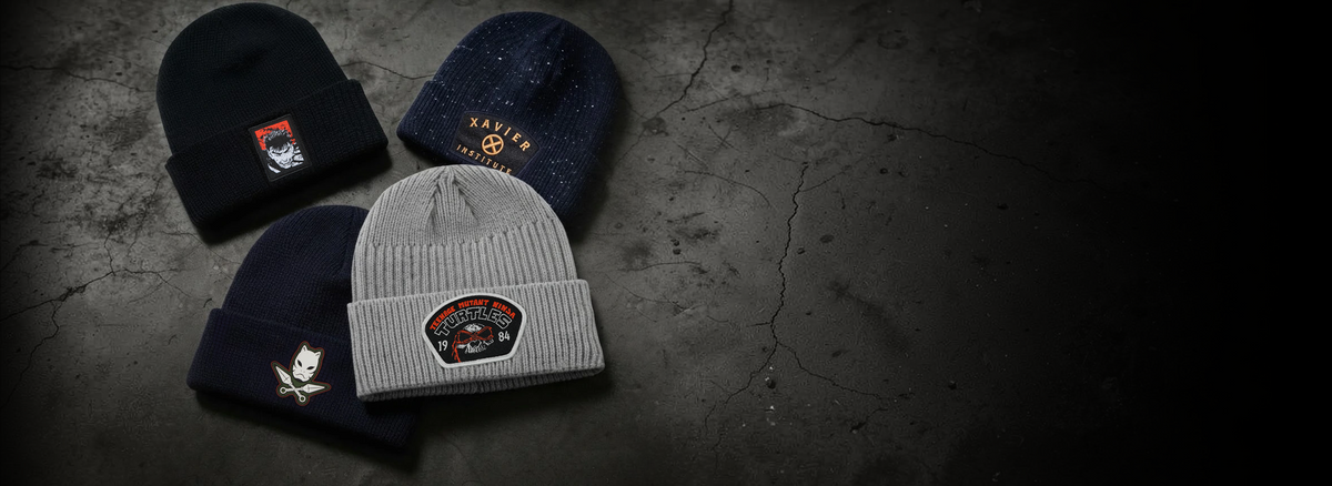 Four knit beanie hats arranged on a dark cracked concrete surface. The hats are black, navy blue, and heather gray, each featuring a custom embroidered logo patch on the front cuff.