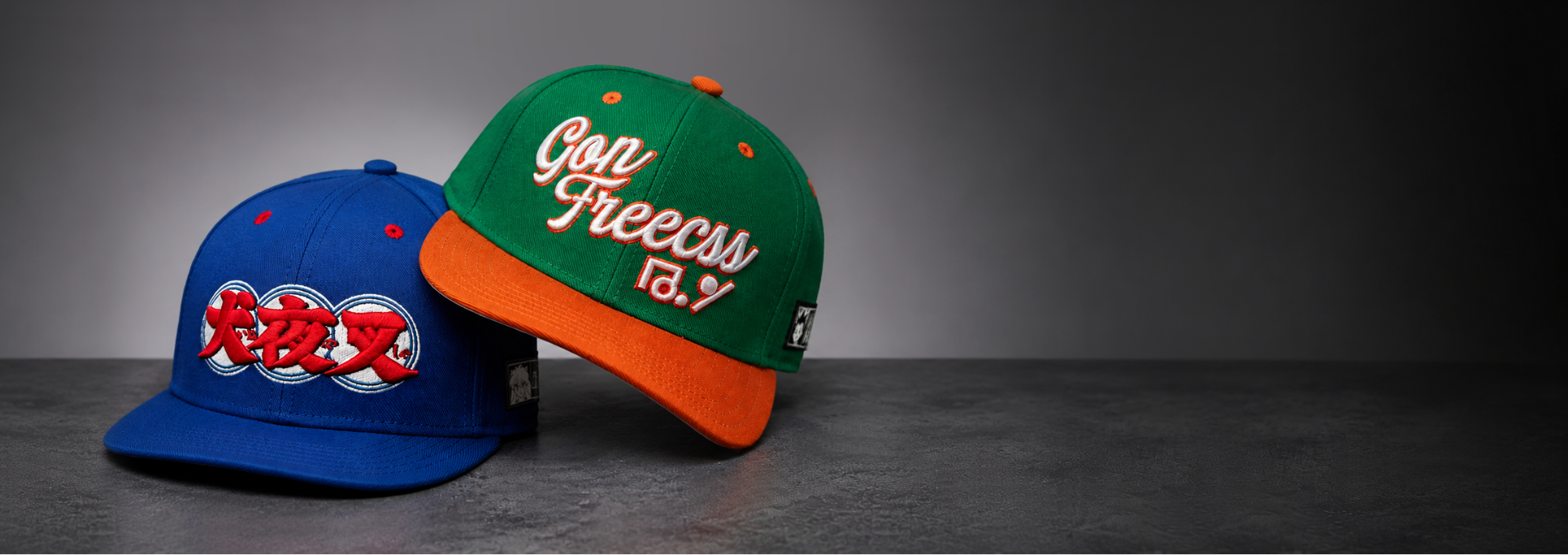 Two snapback caps displayed on a dark gray surface against a dark gray gradient background. The cap on the left is blue with red embroidered characters and a white circular graphic on the front.