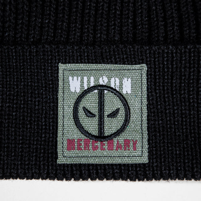 Close-up of a black ribbed knit beanie featuring a square woven patch on the cuff. The patch is sage green with a black circular face emblem, white 'WILSON' text at the top, and pink 'MERCENARY' text at the bottom.