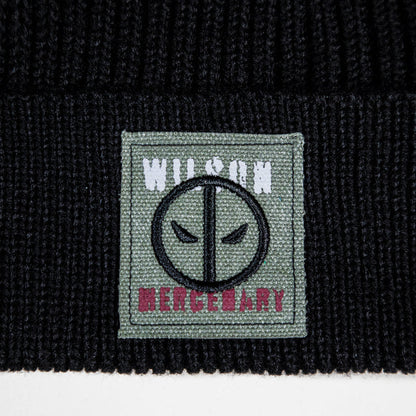 Close-up of a black ribbed knit beanie featuring a square woven patch on the cuff. The patch is sage green with a black circular face emblem, white 'WILSON' text at the top, and pink 'MERCENARY' text at the bottom.