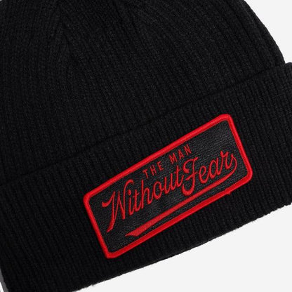 Close-up of a black ribbed knit beanie with a rectangular embroidered patch on the cuff. The patch is black with a red border and red script text reading 'THE MAN Without Fear'.