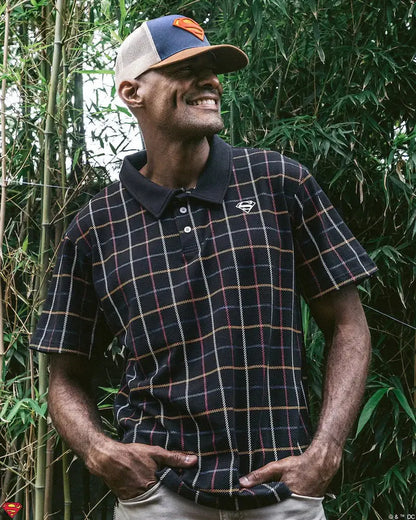 Man wearing a plaid shirt and cap with a dc comics superman logo cap, standing in front of green foliage. available at chimploot.com