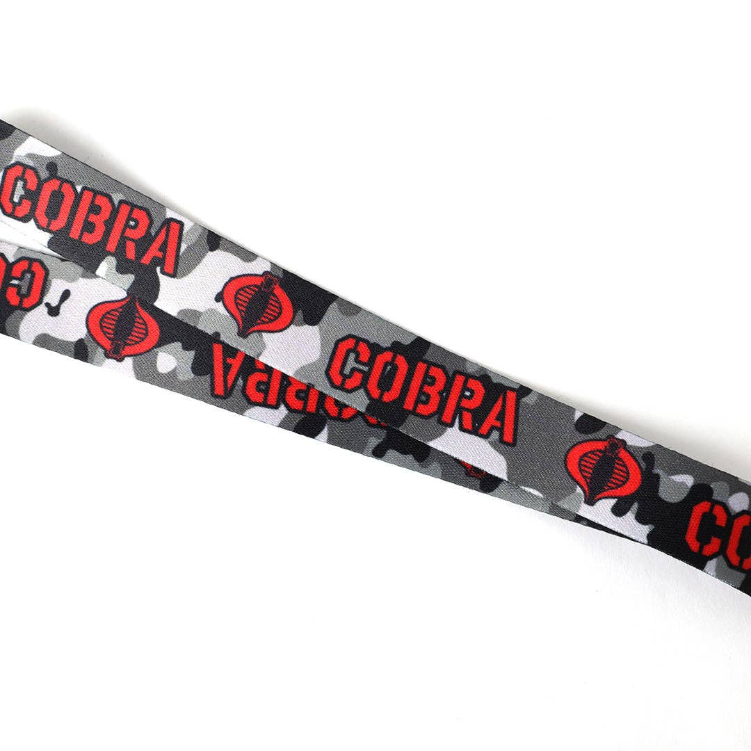 A close-up of a gray camouflage patterned strap featuring red 'COBRA' text and red snake-head logos, photographed on a plain white background.