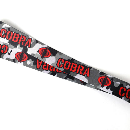 A close-up of a gray camouflage patterned strap featuring red 'COBRA' text and red snake-head logos, photographed on a plain white background.
