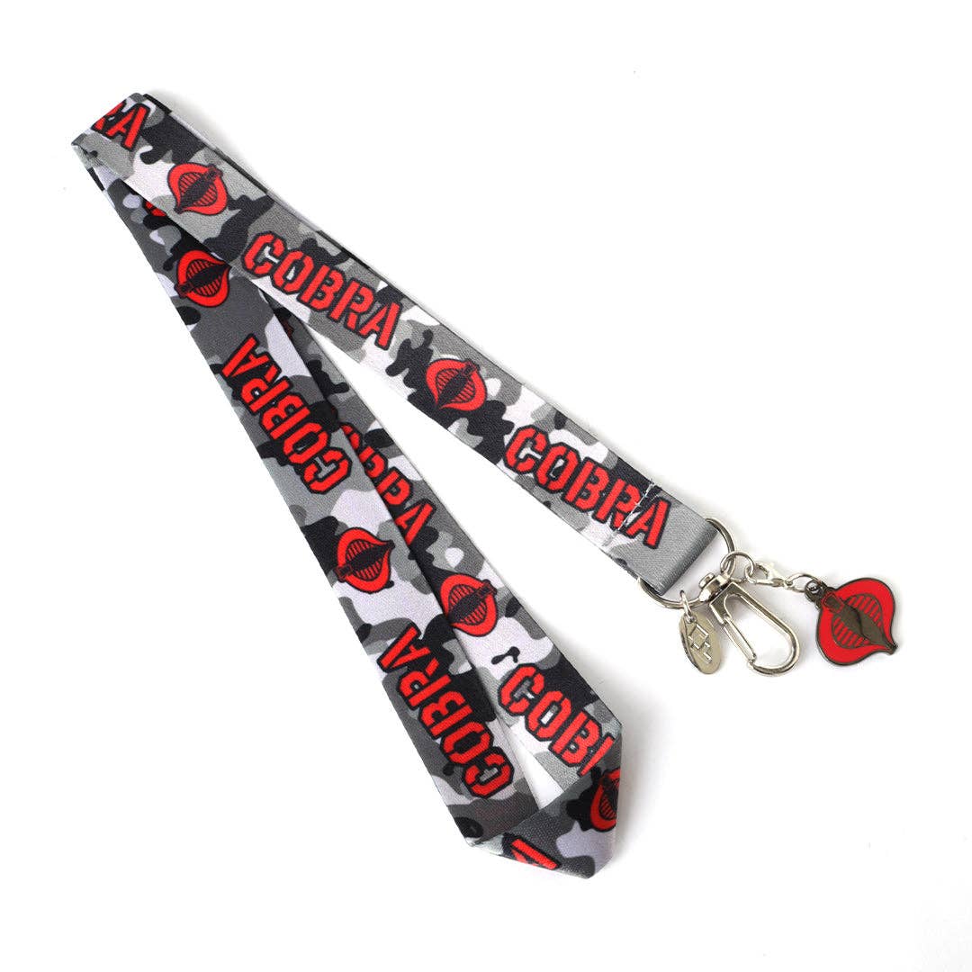 A gray, black, and white camouflage patterned lanyard featuring the word 'COBRA' and a red cobra head logo repeated along the strap, finished with a silver metal lobster clasp and a red-and-black cobra emblem charm, shown on a plain white background.