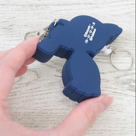 A hand holding a dark blue rubber keychain shaped like a cartoon character, Pokémon Kyogre with silver metal key rings attached, against a light wood-grain background.
