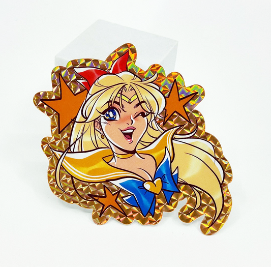 A Sailor Moon enamel lapel pin with a gold-toned holographic border, featuring a blonde character with blue eyes, a red bow, and a blue bow tie, displayed on a clear stand against a white background.
