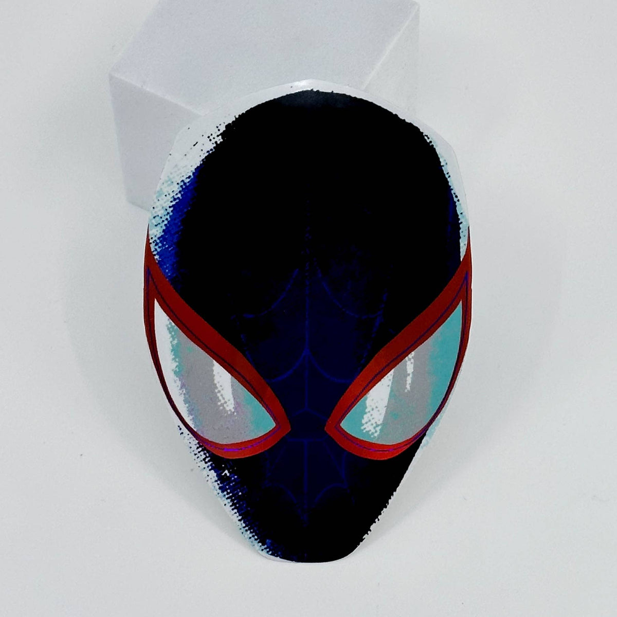 A Spider-Man themed paper mask with a black base, red eye borders, and light blue eyes, displayed on a white block against a white background.